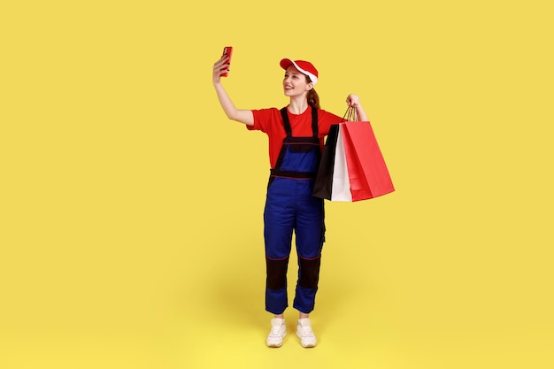 Full length portrait of courier woman standing with shopping bag in hands and taking selfie or having video call with client, wearing overalls and cap. Indoor studio shot isolated on yellow background