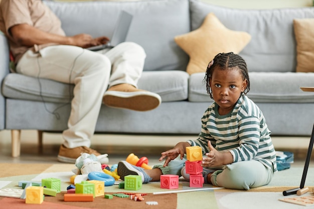 Full length portrait of cute black toddler playing with toy blocks while sitting on floor at home wi