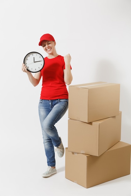 Full length portrait of delivery woman in red cap, t-shirt isolated on white background. Female courier near empty cardboard boxes, holding round clock, showing in time. Receiving package. Copy space.