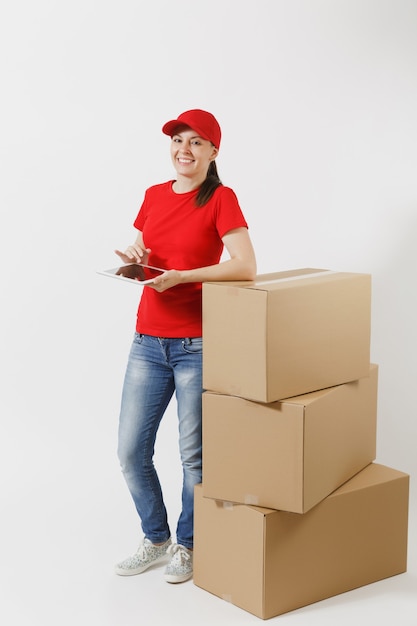 Full length portrait of delivery young woman in red cap, t-shirt isolated on white background. Female courier standing near empty cardboard boxes with tablet pc computer. Receiving package. Copy space