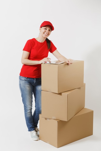Полнометражный портрет молодой женщины доставки в красной кепке, футболке изолированной на белой предпосылке. Женский курьер, стоя возле пустых картонных коробок с планшетным компьютером. Получение пакета. Копировать пространство