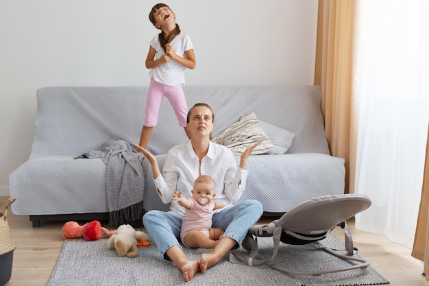 Full length of portrait of depressed mother wearing white shirt and jeans sitting on floor near sofa and feels desperate raised arm dont know what to with screaming children