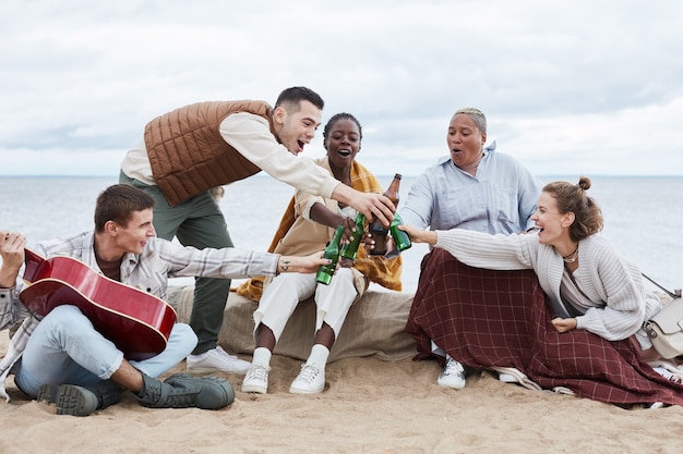 Full length portrait of diverse group of friends on beach in autumn clinking beer bottles and toasting