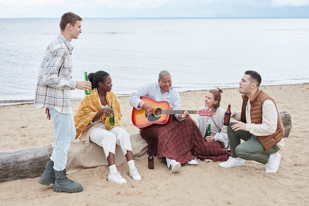 Full length portrait of diverse group of friends enjoying camping on beach in autumn and playing guitar