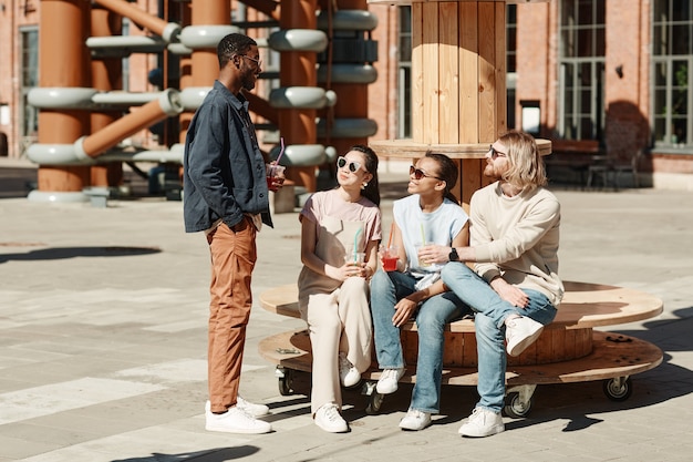 Full length portrait of diverse group of young people chatting while rrelxing in urban city setting