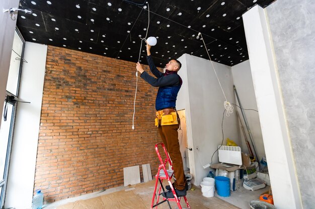 Full Length Portrait Of Electrician On Stepladder Installs Lighting To The Ceiling In Office