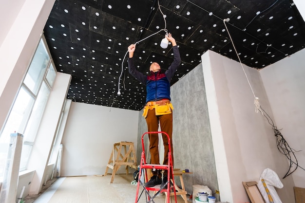 Ritratto a figura intera di elettricista su scala a pioli installa l'illuminazione al soffitto in ufficio