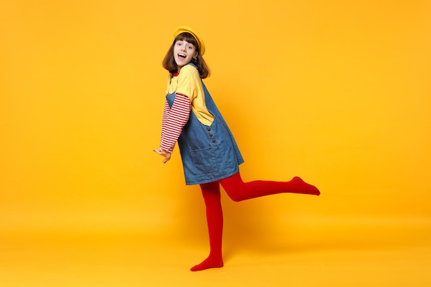 Full length portrait of excited girl teenager in french beret\
and denim sundress standing isolated on yellow wall background in\
studio. people sincere emotions, lifestyle concept. mock up copy\
space.