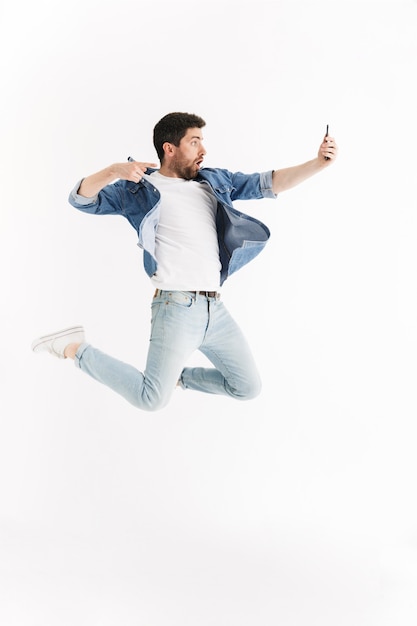 Full length portrait of an excited handsome bearded man wearing casual clothes jumping isolated, taking a selfie
