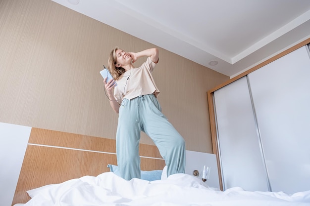 full length portrait of excited happy young female wearing pajama dancing to favorite music on bed in hotel room