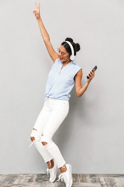 Full length portrait of an excited young woman in sunglasses isolated, holding mobile phone, listening to music with headphones, dancing