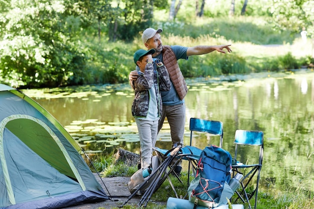 Full length portrait of father and son looking at lake and pointing away while enjoying camping trip together, copy space