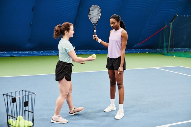 Full length portrait of female tennis coach working with young black woman during practice at indoor