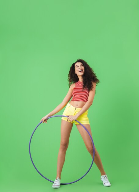 Foto ritratto a figura intera di donna fitness 20s che indossa abiti estivi facendo esercizi con hula hoop durante la ginnastica sul muro verde