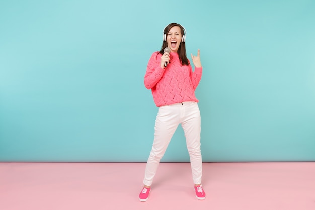 Full length portrait fun woman in knitted sweater, white pants, headphones sing song in microphone 