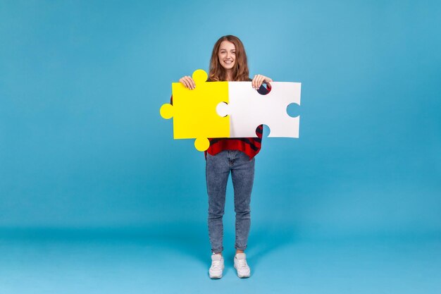 Full length portrait of good looking woman in striped casual sweater, holding big pieces of puzzle, symbol of union and association, solving tasks. Indoor studio shot isolated on blue background.