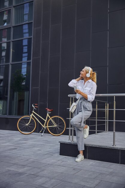 Full length portrait of gorgeous fashionable lady in grey pants and white shirt looking up while listening something in headphones