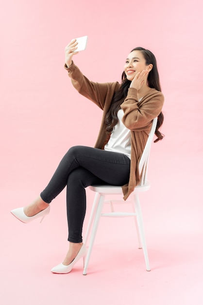Портрет в полный рост великолепной счастливой женщины в деловой одежде, делающей селфи на мобильном телефоне, сидя на стуле