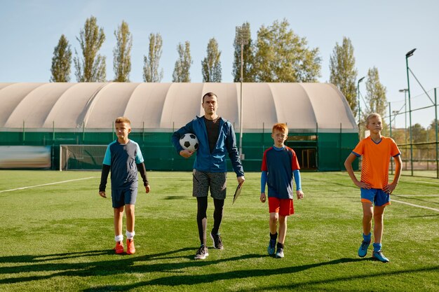 Full-length portrait of handsome coach and junior soccer team walking across field