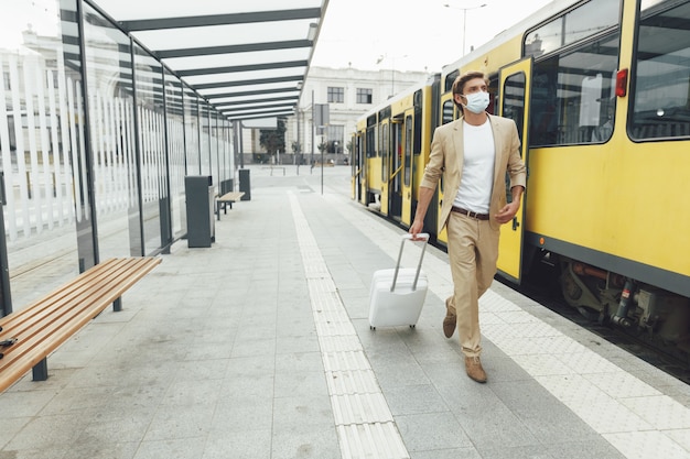 Ritratto integrale dell'uomo bello in vestito di affari e mascherina medica che cammina alla fermata del trasporto pubblico con i bagagli