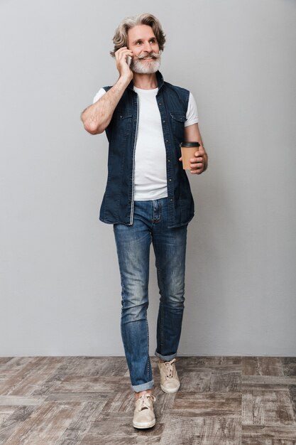 Full length portrait of a handsome smiling stylish mature man wearing a vest standing isolated over gray background, talking on mobile phone while drinking takeaway coffee