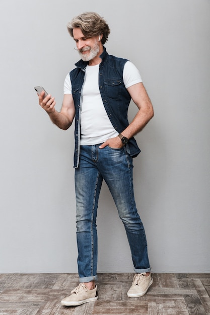 Full length portrait of a handsome smiling stylish mature man wearing a vest walking over gray background, using mobile phone