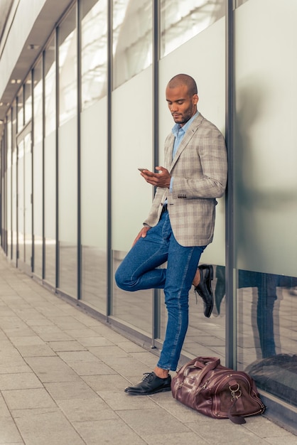彼の茶色の革のバッグの近くに屋外で立っているスマートフォンを使用してハンサムなスタイリッシュな若いアフロアメリカ人実業家の全身像
