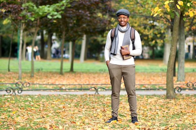 Full length portrait of happy black college or university student guy, young African Afro American man in glasses, hat, scarf with backpack and book is walking in golden autumn park at fall, smiling