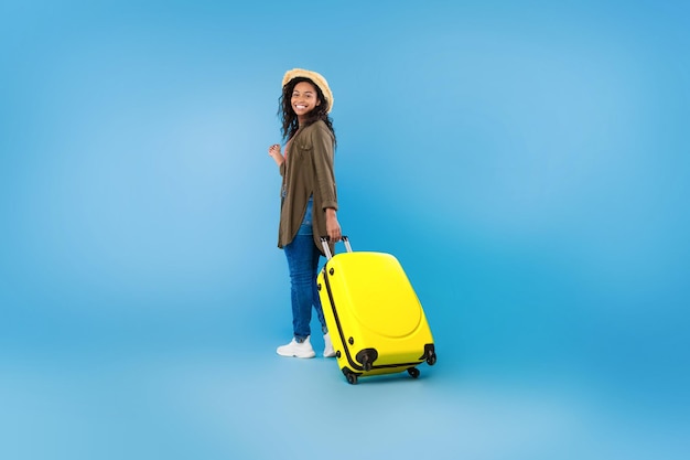 Full length portrait of happy black female traveler walking with suitcase over blue studio