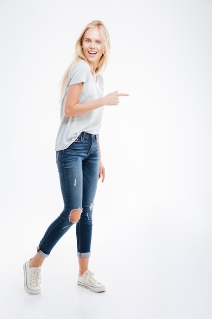 Full length portrait of a happy casual woman pointing finger away isolated on a white wall