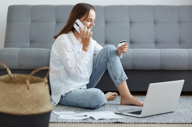 Full length portrait of happy exited woman wearing white shirt and jeans sitting on floor near sofa using laptop and talking phone holding credit card and expressing happiness