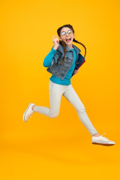 Full length portrait of happy little schoolgirl jumping on yellow background teen girl on school break childhood happiness finally graduated movement and people concept crazy kid jumping in air