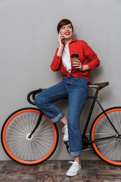 회색 벽에 격리된 자전거 옆에 서 있는 동안 스마트폰으로 이야기하고 커피 컵을 들고 있는 행복한 여성의 전체 길이 초상화