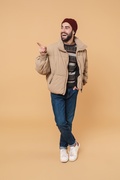 Full length portrait of a happy young bearded man wearing winter jacket standing isolated on beige, pointing finger away at copy space