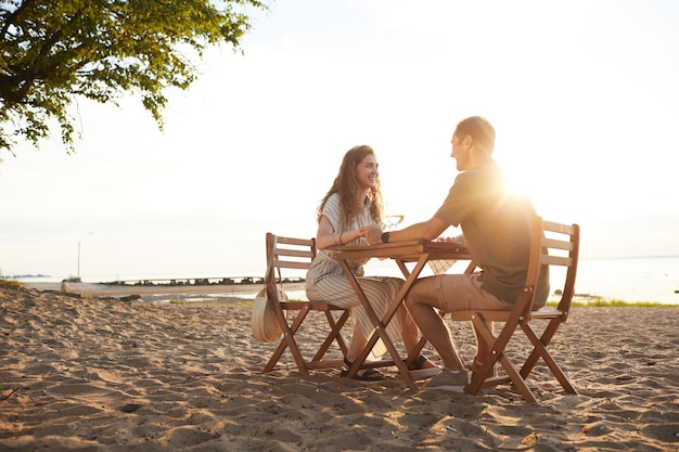 Full length portrait of happy young couple drinking wine at picnic outdoors while enjoying romantic ...