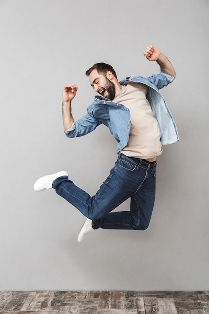 Full length portrait of a happy young man wearing shirt jumping over gray wall, celebrating