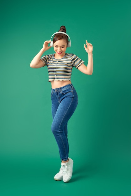 Full length portrait of joyful Asian woman on casual clothing dancing and listening to music with headphone over green background