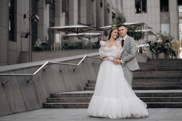 Full length portrait of just married couple posing outdoors Bride standing with closed eyes smiling groom huggs bride Concept of love and marriage