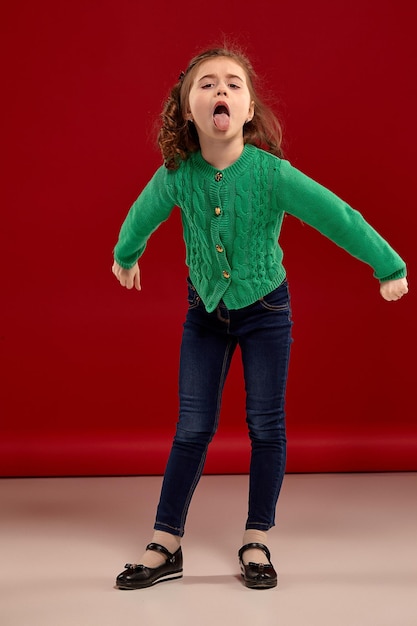Full length portrait of a little brunette girl with a long curly hair posing against a red background