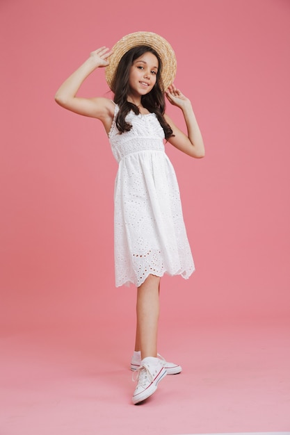 Full length portrait of lovely summer girl 8-10 wearing white dress posing at camera touching straw hat with smile, isolated over pink background