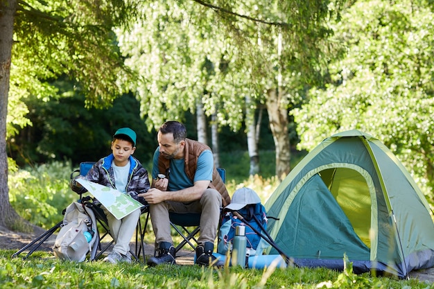 Full length portrait of loving father and son looking at map while enjoying camping trip together in nature, copy space