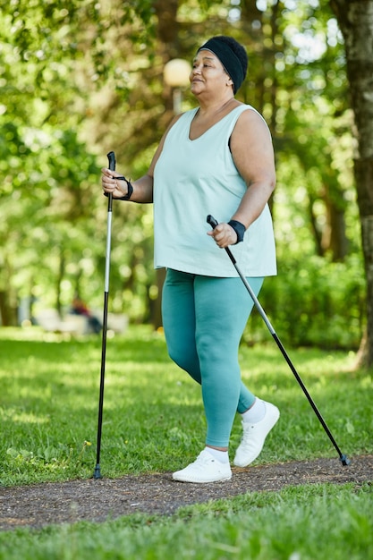 Full length portrait of mature black woman walking with poles in park during outdoor workout