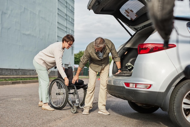 Full length portrait of mature couple unloading wheelchair out
of car trunk in parking lot outdoors ...