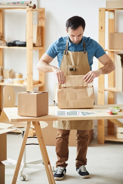 Full length portrait of mature man wearing apron packaging orders while standing by wooden table, food delivery service worker