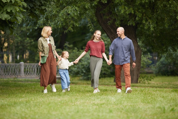 Full length Portrait of modern carefree family with two kids holding hands while walking on green grass outdoors