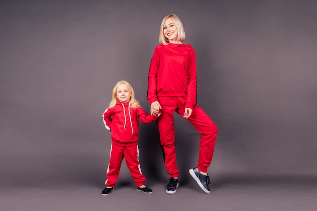 Full length portrait mother with the daughter in sports suits.beautiful young blonde woman with a little girl posing in red stylish suit studio black background,active sporting family hip-hop dancer.