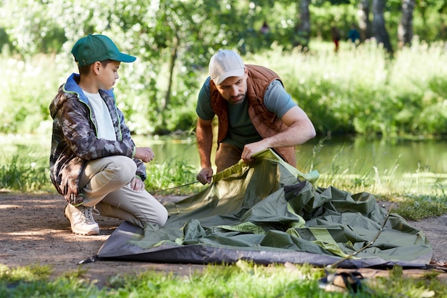 Фото Портрет в полный рост отца и сына, устанавливающих палатку вместе во время кемпинга у озера в лесу, копия пространства