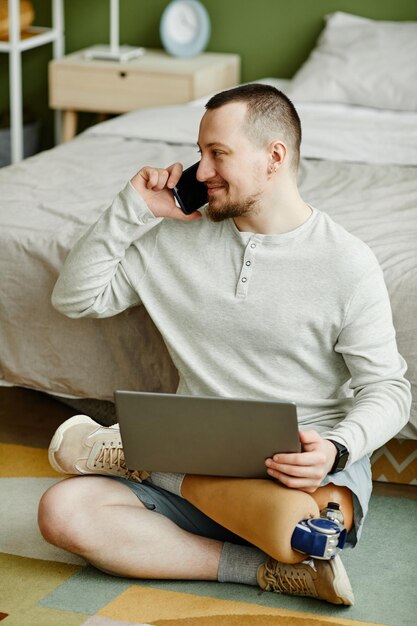写真 家の床に座りながらノートパソコンを使って義足を持った笑顔の男性の全長のポートレート