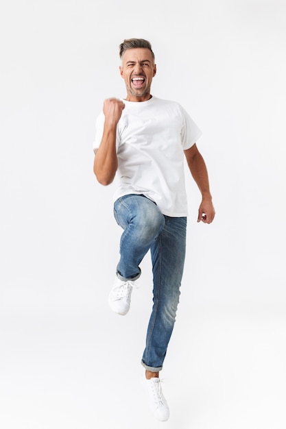 Ritratto a figura intera di un uomo positivo di 30 anni che indossa una maglietta casual e jeans che stringono i pugni isolati su bianco