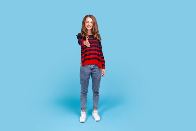 Full length portrait of positive pleasant woman wearing striped casual style sweater giving hand to handshake and smiling friendly meet new people Indoor studio shot isolated on blue background